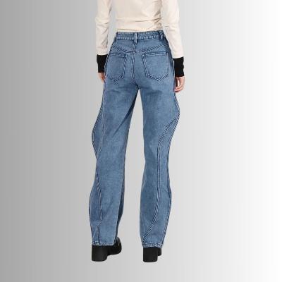 Vrouw in blauwe jeans met unieke golvende naden, staand tegen een grijze achtergrond. Modieuze damesbroek, casual stijl, trendy denim.