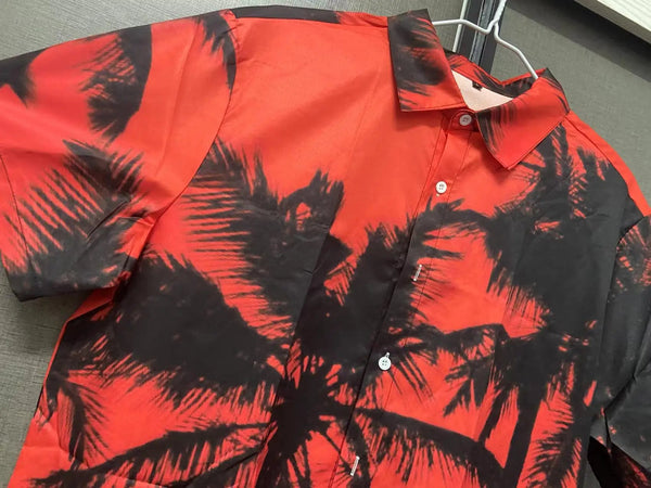 Rode Hawaii-overhemd met zwarte palmboomprint, korte mouwen en knoopsluiting, hangend aan een kledinghanger. Perfect voor zomerse casual outfits.