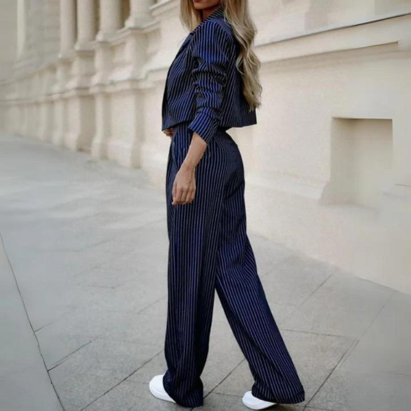 Vrouw in blauw gestreept pak met lange broek en jasje, witte sneakers, staand op trottoir voor klassieke architectuur. Modieuze dameskleding.