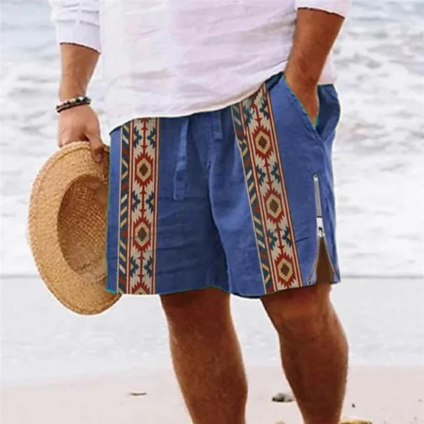 Man in blauwe strandshort met etnisch patroon en rits, houdt strohoed vast op zandstrand. Zomermode, strandkleding, herenkleding.