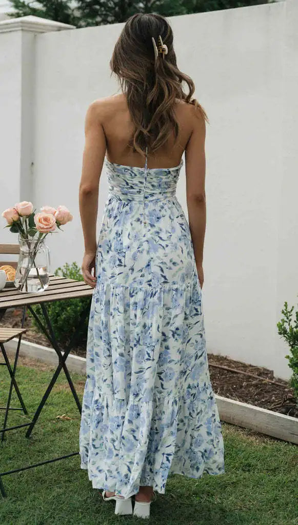 Vrouw in lange, blauwe bloemenjurk met blote rug, staand in tuin naast tafel met rozen. Zomermode, elegante dameskleding, buitenstijl.