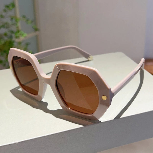 Zonnebril met zeshoekige, beige montuur en bruine glazen op een grijze achtergrond. Modieuze damesaccessoire, perfect voor zonnige dagen.