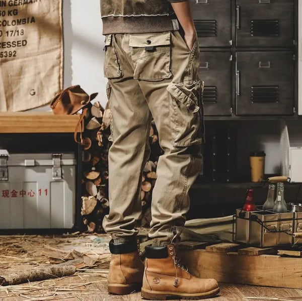 Man in beige cargo pants en bruine laarzen, staand in een industriële ruimte met houtblokken en metalen kisten. Robuuste werkkleding, casual stijl.