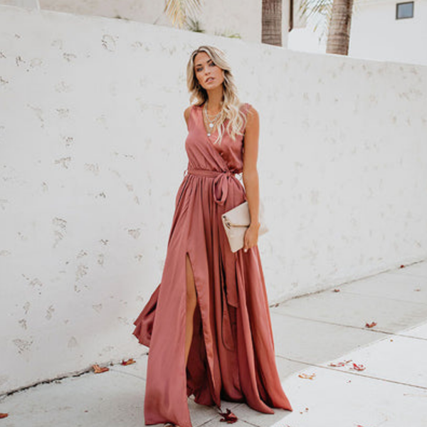 Jonge vrouw in elegante roze avondjurk met hoge split, poseert buiten tegen witte muur. Perfect voor mode, zomerjurken, avondkleding.