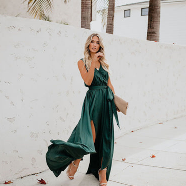 Vrouw in elegante groene avondjurk met hoge split, wandelend langs witte muur. Perfect voor mode, zomerjurken, avondkleding.