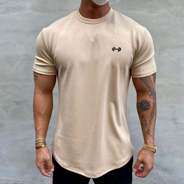 Man in beige T-shirt met halterlogo, toont gespierde armen en tatoeages, draagt gouden armbanden en horloge, tegen grijze achtergrond.