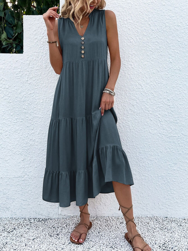 Vrouw in elegante, mouwloze, grijze zomerjurk met knopen, staand tegen witte muur. Perfecte outfit voor casual zomermode.