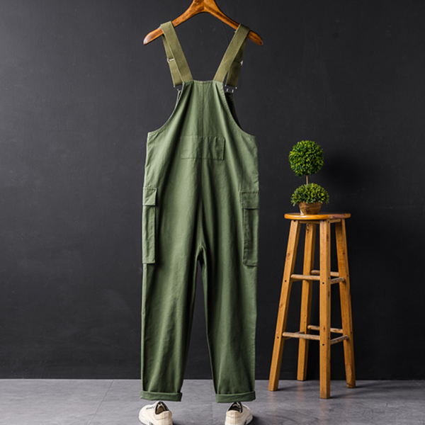 Groene unisex tuinbroek op houten hanger, naast houten kruk met decoratieve plant, tegen zwarte achtergrond. Comfortabele werkkleding, modetrend.