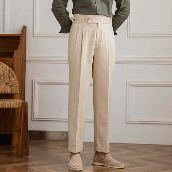 Beige linnen broek voor heren, hoge taille, geplooid ontwerp, casual stijl, gecombineerd met lichtbruine loafers, houten vloer en witte muur.