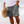 Man in grijze korte broek met palmboomprint, strandachtergrond, zomermode, casual herenkleding, strandhoed in hand, ontspannen sfeer.
