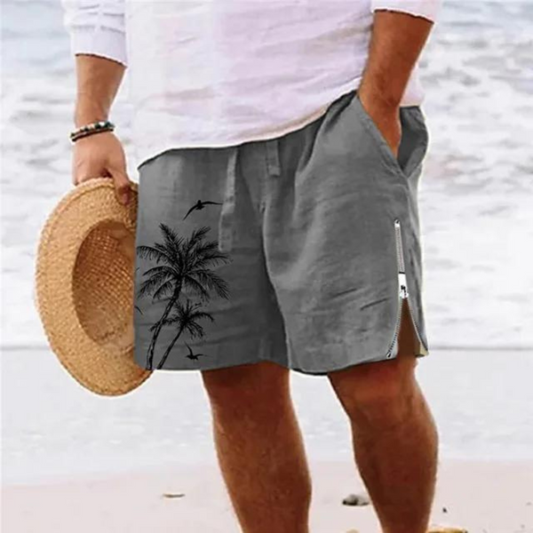 Man in grijze korte broek met palmboomprint, strandachtergrond, zomermode, casual herenkleding, strandhoed in hand, ontspannen sfeer.