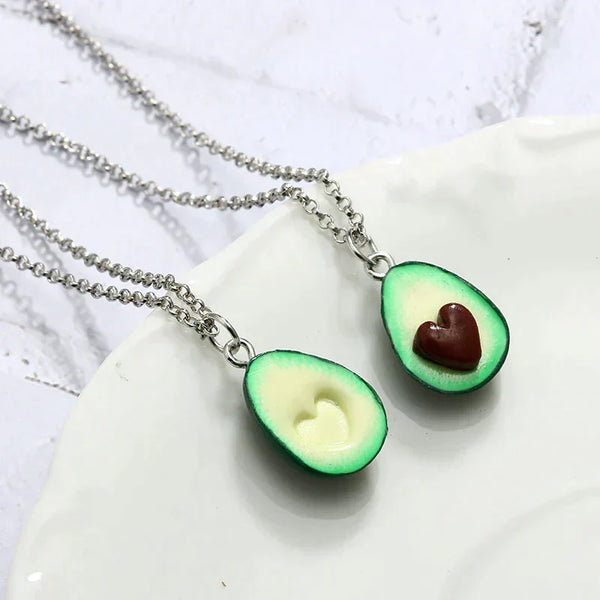 Twee zilveren kettingen met avocado-hangers, elk met een hartvormige uitsparing, op een witte achtergrond. Perfect als vriendschapskettingen.