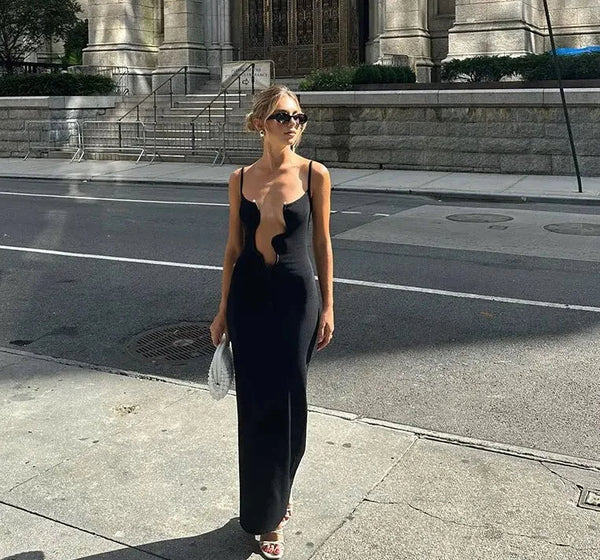 Vrouw in elegante zwarte avondjurk met diepe halslijn, draagt zonnebril en witte handtas, loopt op zonnige stadsstraat. Fashion, stijl, mode.