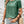 Vrouw in groene T-shirt en denim shorts met rieten tas, sieraden en golvend haar. Casual zomeroutfit, modetrend, dameskleding.