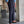 Man in blauwe blouse en donkergrijze pantalon met bruine loafers, staand naast betonnen muur en plant. Stijlvolle herenmode, zakelijke outfit.