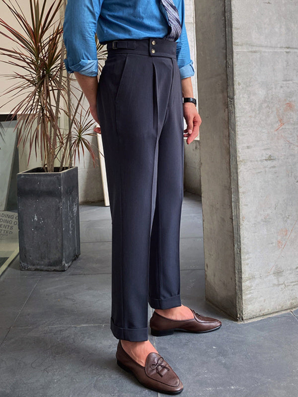 Man in blauwe blouse en donkergrijze pantalon met bruine loafers, staand naast betonnen muur en plant. Stijlvolle herenmode, zakelijke outfit.