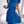Vrouw in elegante blauwe plisséjurk met strikdetail, gouden kettingtas, staand voor witte bank. Modieuze dameskleding, zomerjurk.