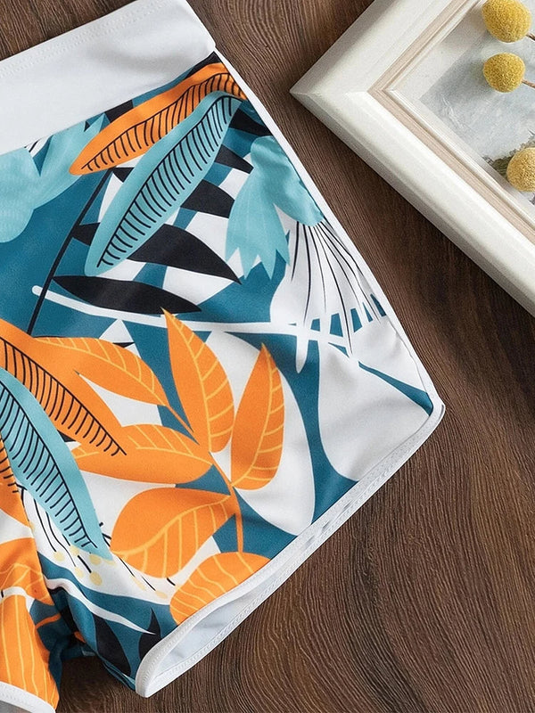 Kleurrijke zwembroek met tropisch bladpatroon op houten achtergrond, naast een ingelijste foto. Perfect voor strandmode en zomerse stijl.
