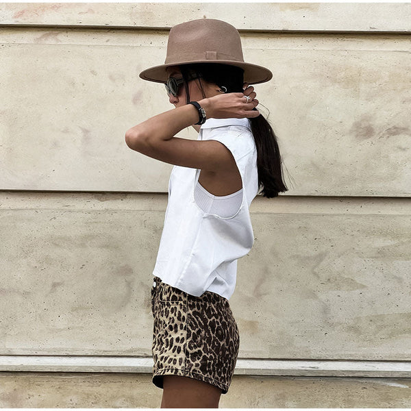 Vrouw in witte mouwloze top, luipaardprint shorts en bruine hoed, tegen beige muur. Zomermode, casual stijl, trendy outfit.