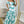 Vrouw in elegante, off-shoulder maxi-jurk met groen-wit bloemenpatroon, staand voor een grote spiegel in een stijlvolle kamer.