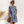 Vrouw in gestreepte zomerjurk met blauwe en witte strepen, draagt zonnebril en houdt een rotan handtas vast. Perfecte outfit voor zomermode.