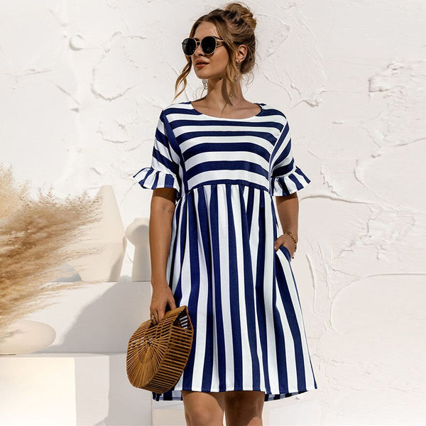 Vrouw in gestreepte zomerjurk met blauwe en witte strepen, draagt zonnebril en houdt een rotan handtas vast. Perfecte outfit voor zomermode.