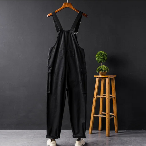 Zwarte tuinbroek op houten hanger tegen donkere muur, naast houten kruk met groene plant. Modieuze unisex kleding, casual stijl.