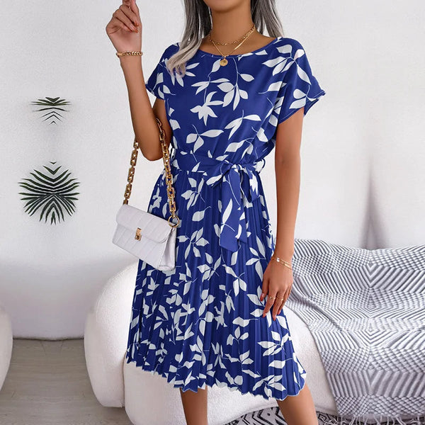 Blauwe zomerjurk met witte bloemenprint, korte mouwen en ceintuur, gedragen door vrouw met witte schoudertas. Perfect voor casual en elegante gelegenheden.