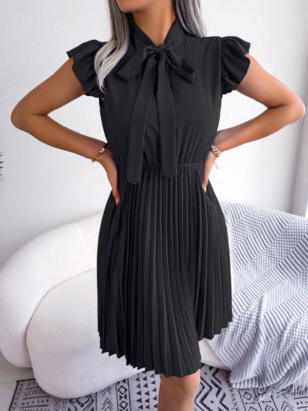Vrouw in zwarte geplooide jurk met strik, staand voor witte bank. Elegante damesmode, zomerjurk, casual chic outfit, trendy kledingstijl.