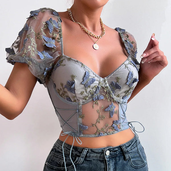 Vrouw in doorschijnende blouse met bloemenborduursel en pofmouwen, gecombineerd met een gouden ketting en spijkerbroek. Modieuze dameskleding.