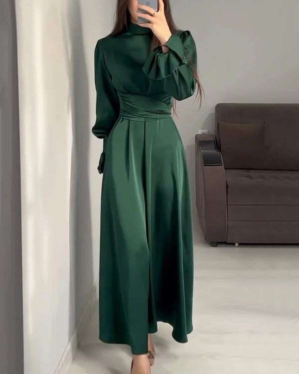 Vrouw in elegante groene satijnen jurk met lange mouwen, staand in moderne woonkamer. Perfect voor avondkleding of formele gelegenheden.