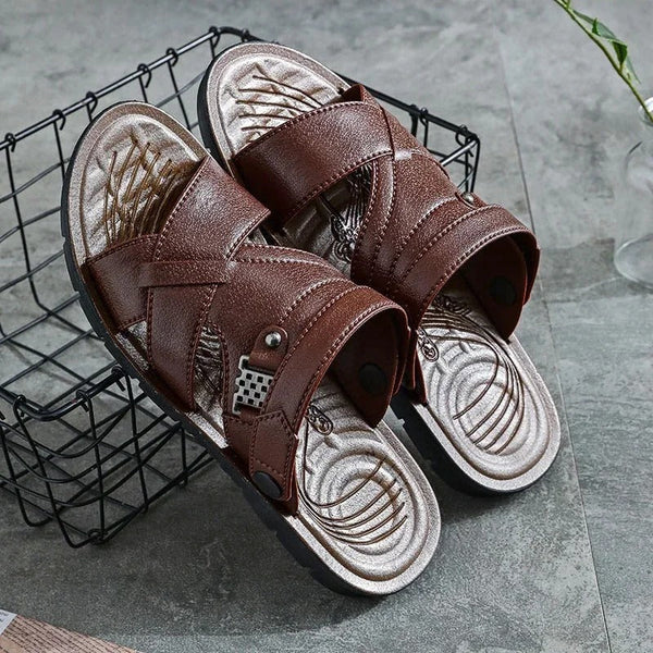 Bruine leren sandalen met kruisbanden en metalen gesp, op een grijze achtergrond. Comfortabele herenslippers, ideaal voor casual zomeroutfits.