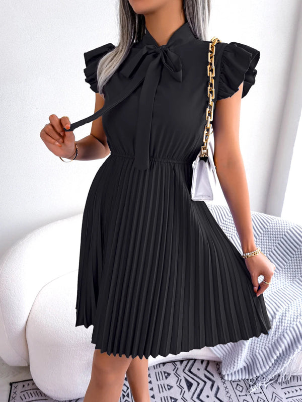 Vrouw in elegante zwarte plissé jurk met strik en ruches, gouden kettingtas, staand in moderne woonkamer. Perfect voor mode en stijl inspiratie.