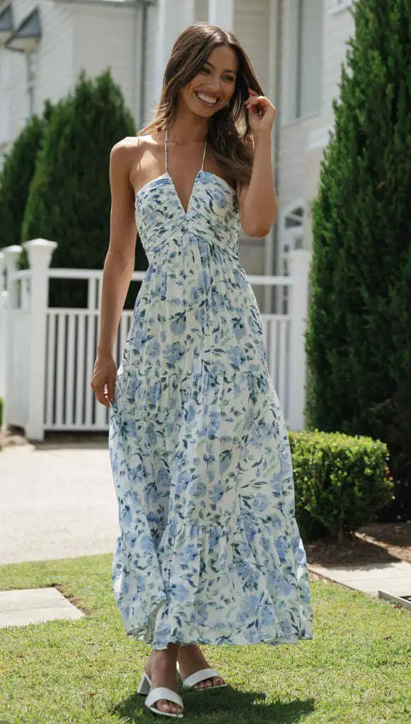 Vrouw in lange, zomerse bloemenjurk met halternek, wandelend buiten bij witte hekken en groene struiken, modieuze casual outfit.