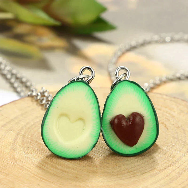 Twee avocado-hangerkettingen met hartvormige pit, liggend op houten ondergrond. Perfecte sieraden voor avocado-liefhebbers en vriendschapskettingen.