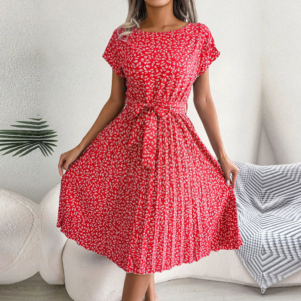 Vrouw in rode bloemenjurk met korte mouwen en strikceintuur, staand in moderne woonkamer. Zomerse damesmode, casual elegantie.