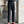 Man in blauwe blouse en zwarte hoge taille broek met bruine loafers, staand naast betonnen pilaar en plant. Stijlvolle herenmode, zakelijke outfit.