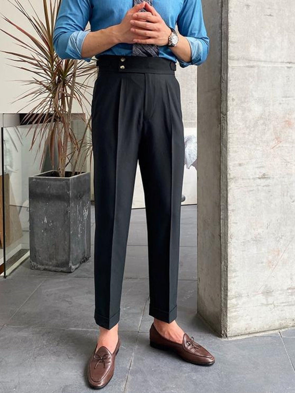 Man in blauwe blouse en zwarte hoge taille broek met bruine loafers, staand naast betonnen pilaar en plant. Stijlvolle herenmode, zakelijke outfit.