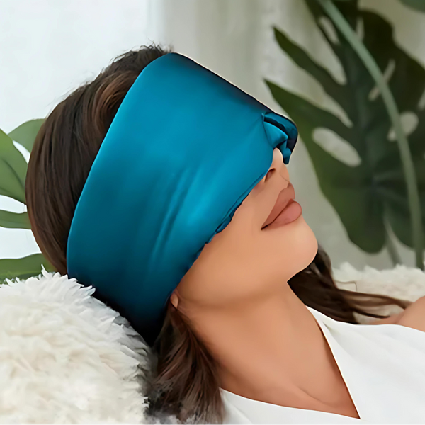 Vrouw ontspant met blauwe zijden slaapmasker, liggend op witte kussens. Luxe slaapaccessoire, ideaal voor rust en comfort.