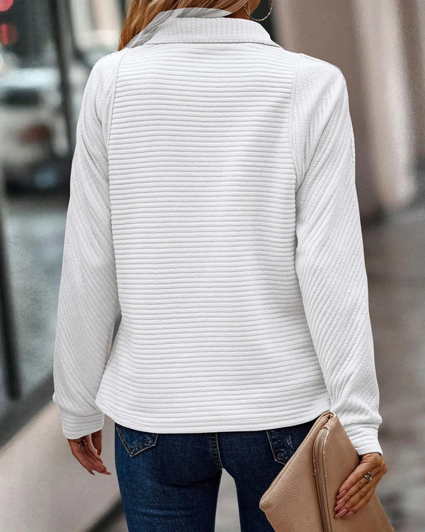 Vrouw in witte geribbelde trui en blauwe jeans, houdt beige clutch vast. Modieuze herfstoutfit, casual straatstijl, stedelijke achtergrond.