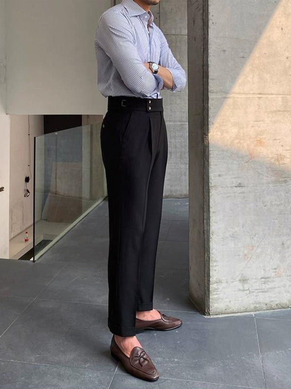 Man in stijlvolle zakelijke outfit met gestreept overhemd, zwarte pantalon en bruine loafers, leunend tegen een betonnen muur in moderne kantooromgeving.