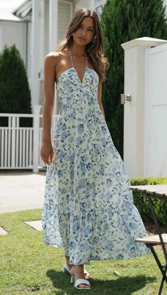Vrouw in lange, blauwe bloemenjurk met halternek, wandelend in tuin. Zomerse mode, elegante dameskleding, casual chique stijl.