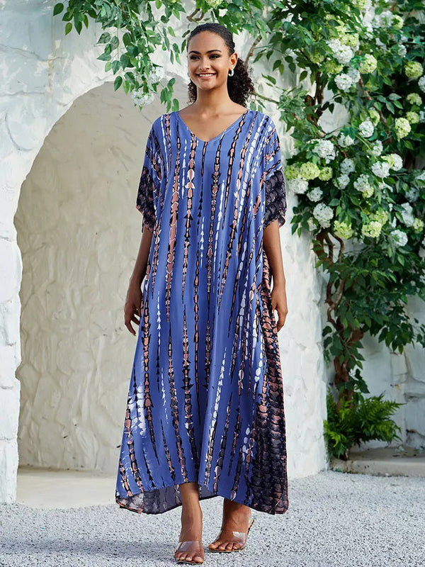 Vrouw in blauwe maxi-jurk met abstract patroon, staand voor witte stenen muur en groene planten. Zomermode, elegante dameskleding.