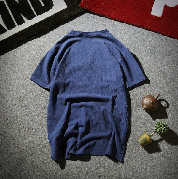 Blauwe katoenen blouse met korte mouwen op grijze vloer, naast een theepot en plant. Casual damesmode, comfortabele zomerstijl.