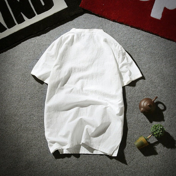 Witte katoenen blouse met korte mouwen op grijze vloer, naast een theepot en kleine plant. Casual kleding, minimalistisch design.