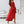 Vrouw in elegante rode off-shoulder jurk met ruches, staand in moderne witte woonkamer. Trendy zomeroutfit, stijlvolle mode, chique kleding.
