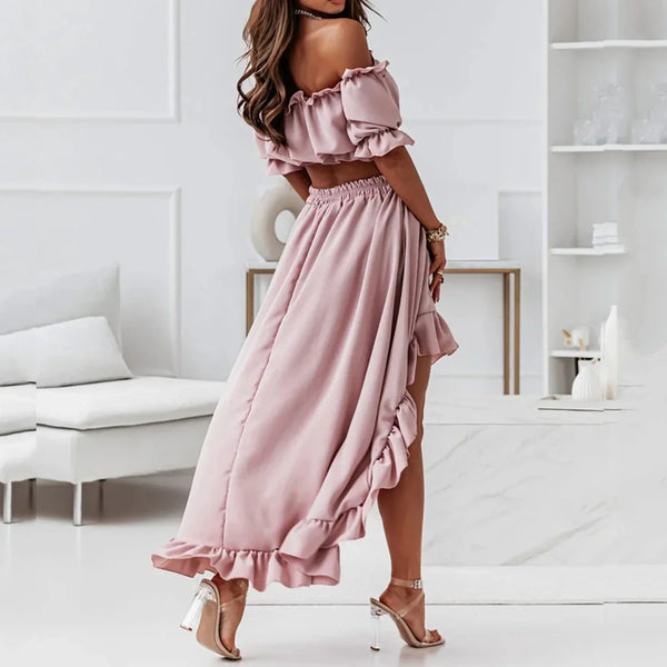 Vrouw in roze off-shoulder jurk met ruches, staand in moderne witte woonkamer. Elegante zomeroutfit, hoge hakken, stijlvolle mode.