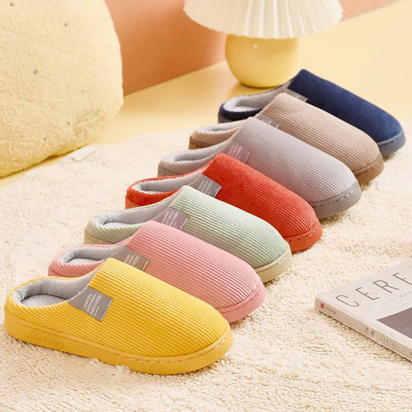 Zeven paar kleurrijke ribfluwelen pantoffels op een beige tapijt, naast een boek en lamp. Perfect voor comfortabele, stijlvolle binnenhuismode.