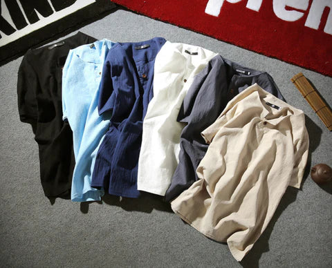 Zes casual T-shirts in verschillende kleuren (zwart, lichtblauw, donkerblauw, wit, grijs, beige) liggen op een rij op een grijze vloer.