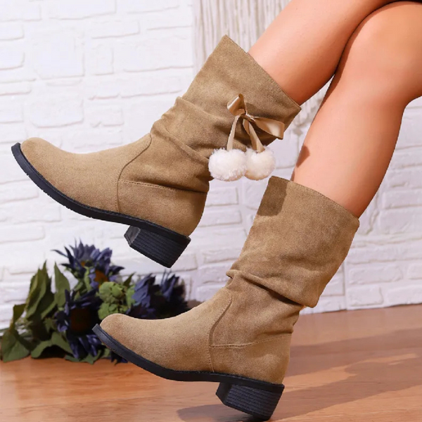 Beige suède laarzen met pompons, lage hak en ronde neus, gedragen door model tegen witte bakstenen muur. Trendy damesmode, herfst/winter schoenen.